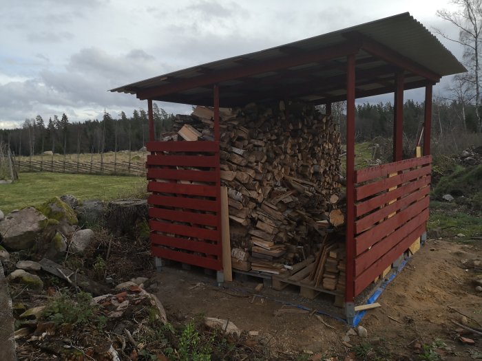 Ett hembyggt vedskjul med röda väggar och en blå presenning på marken, fullt med staplad ved.