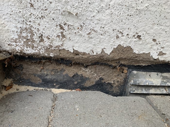 Skadad husfasad med flagnande färg och spruckna puts vid sockeln, synligt nedre delen av en vitmålad vägg och trottoaren.
