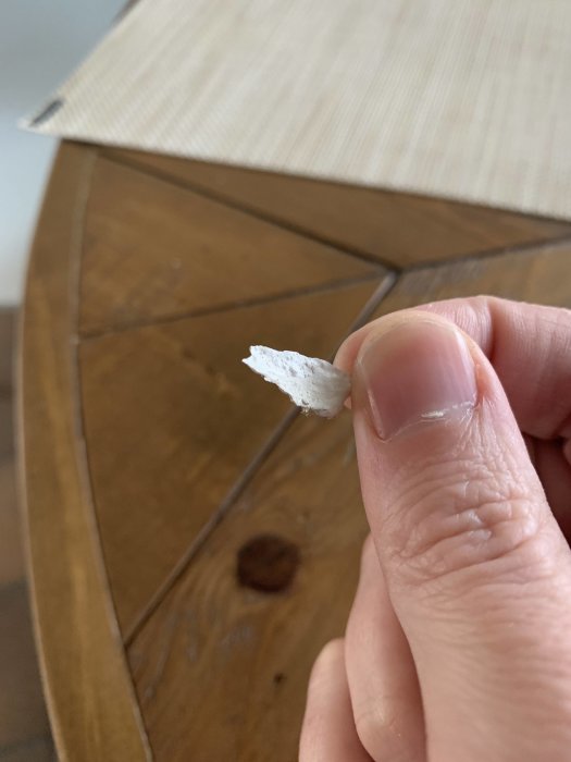 Närbild på hand som håller en bit avsläppt vit kalkfärg framför en brunt träunderlag.