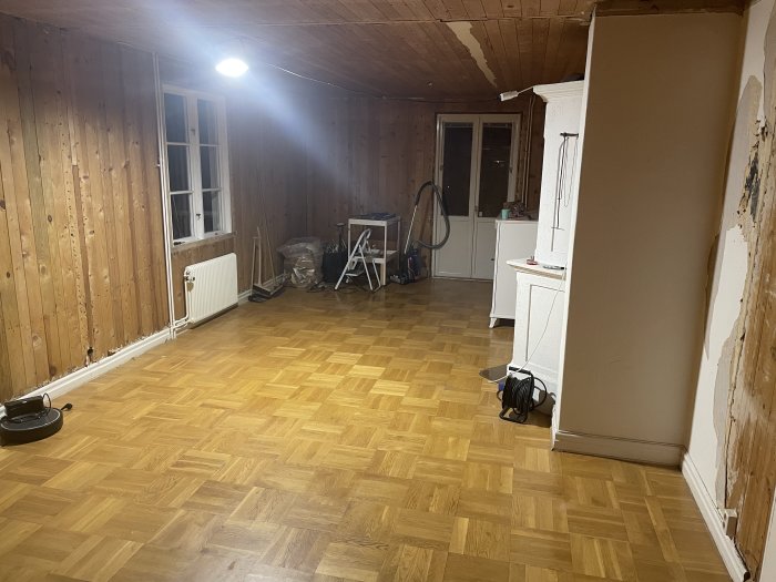 Rum under renovering med träpanelväggar, parkettgolv och en vit kakelugn.