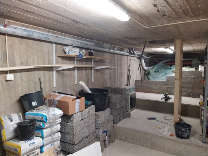 Bild på en källare under renovering med byggmaterial, en avfuktare och en ventilationsslang intill en trappa.