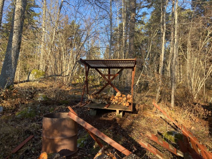 Ett vedskjul fyllt med vedstockar under ett tunt tak omgivet av skog och rörlig mark.