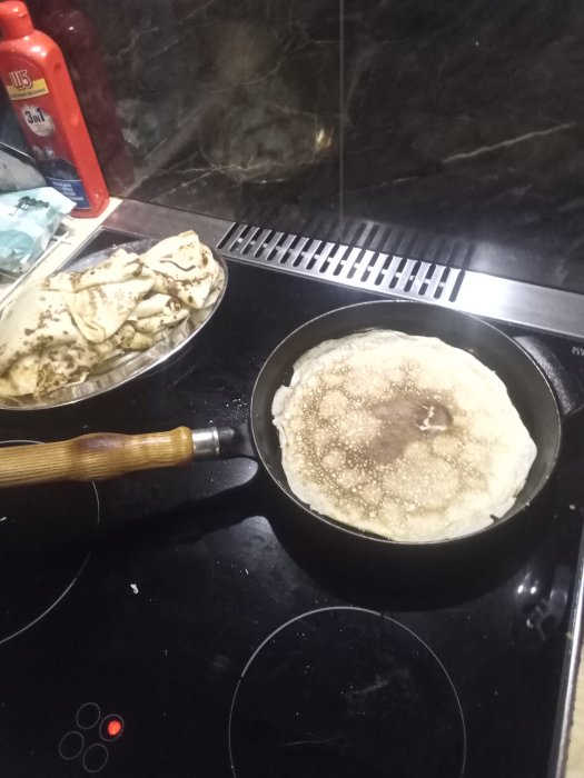 Pannkakor på spisen, en stekning och en hög med färdiga, på svart spishäll.