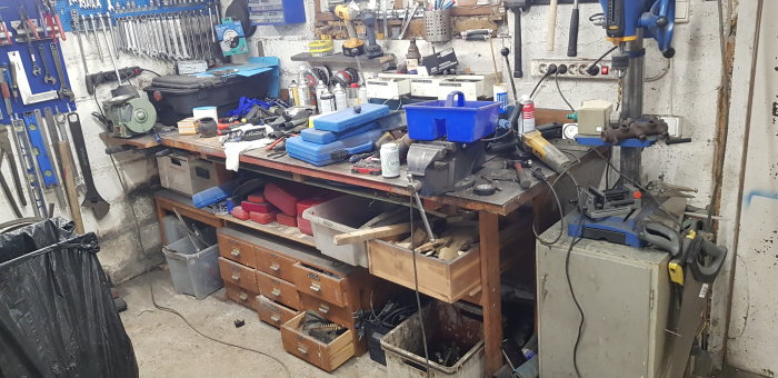 Arbetsbänk i garage överfylld med verktyg och delar, vilket ger ett rörigt intryck.