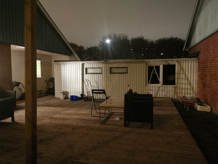 Kvällsbild av en bakgård med träterrass, möbler och ett vitmålat förråd/garage med en regnränna längs taket.