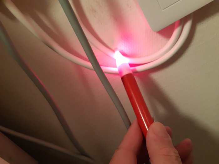 Hand som håller en spänningsprovare mot en vägg med synliga elektriska kablar i olika färger.