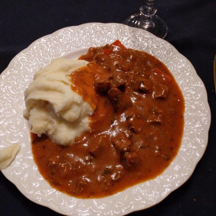 Gulaschgryta med röd sås och köttbitar serverad med potatismos på ett vitt dekorerat fat.