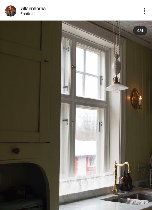 Kök med fönster över diskbänk med cafégardin, kopparmatur och hängande lampa.