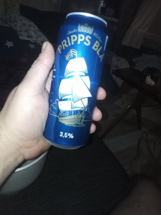 Hand håller en blå ölburk med texten 'PRIIPPS BLÅ' i ett mörkt rum, avslappnad atmosfär.