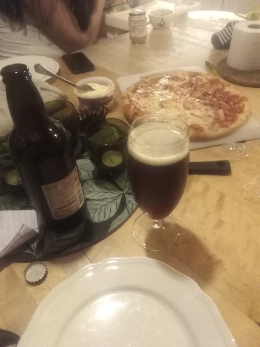 En julölflaska och ett fyllt glas bredvid en pizza på ett köksbord, med soft bakgrundsbelysning.