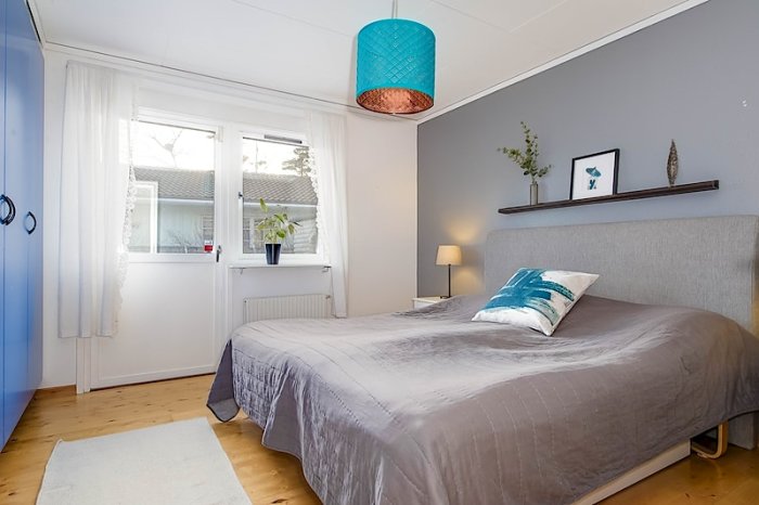 Renoverat sovrum med grått väggfärg, en dubbelsäng med grått överkast, vita gardiner och en blå taklampa.