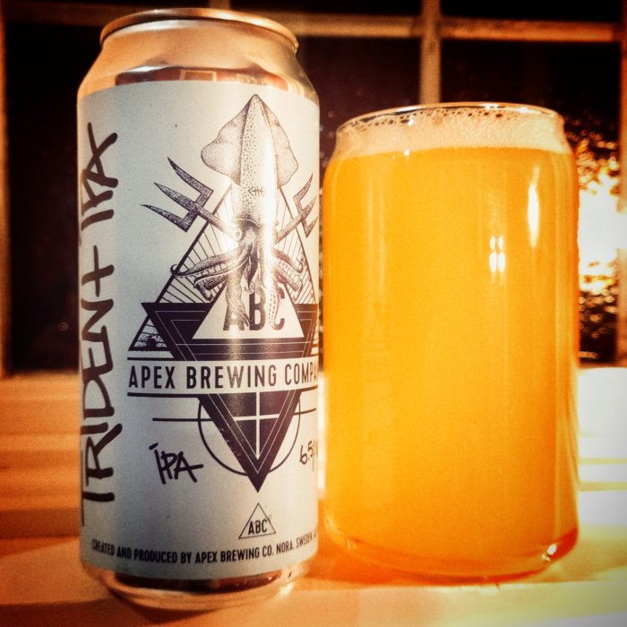 Burk av Apex Brewing Company öl med bläckfiskdesign bredvid ett glas fyllt med orangefärgad IPA.