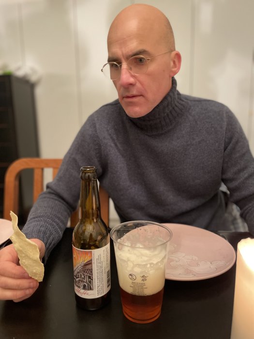 Man som håller en samosa med en flaska öl och ett glas fylld med ale på bord framför honom.