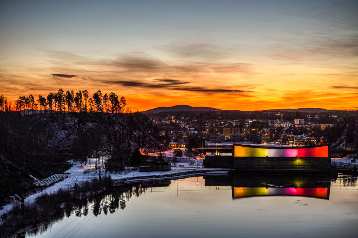 Solnedgång över stad med färgglad byggnad speglad i vattendrag och silhuett av träd på kulle.