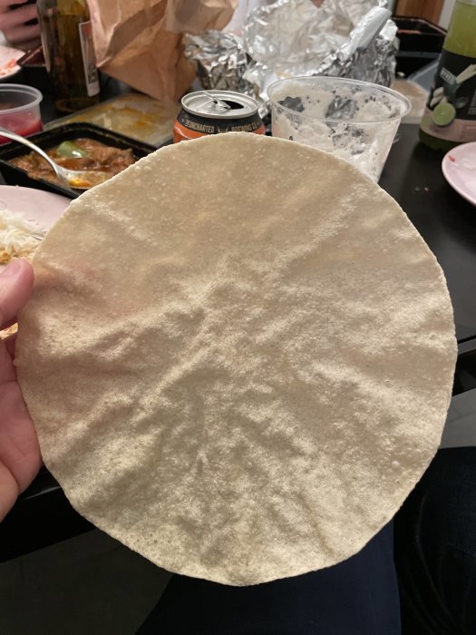 En person håller ett stort rundat pappadambröd framför ett matbord med diverse matrester och en burk öl.