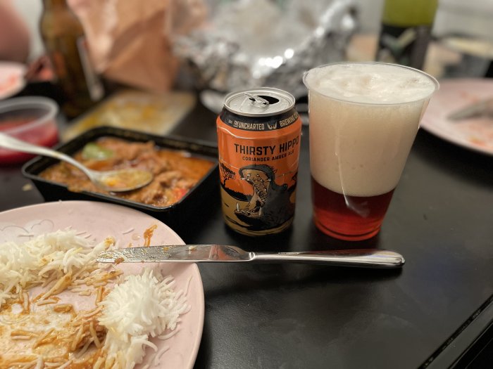 Burk med Thirsty Hippo öl bredvid ett glas med öl och skum, matrester på en tallrik i förgrunden.