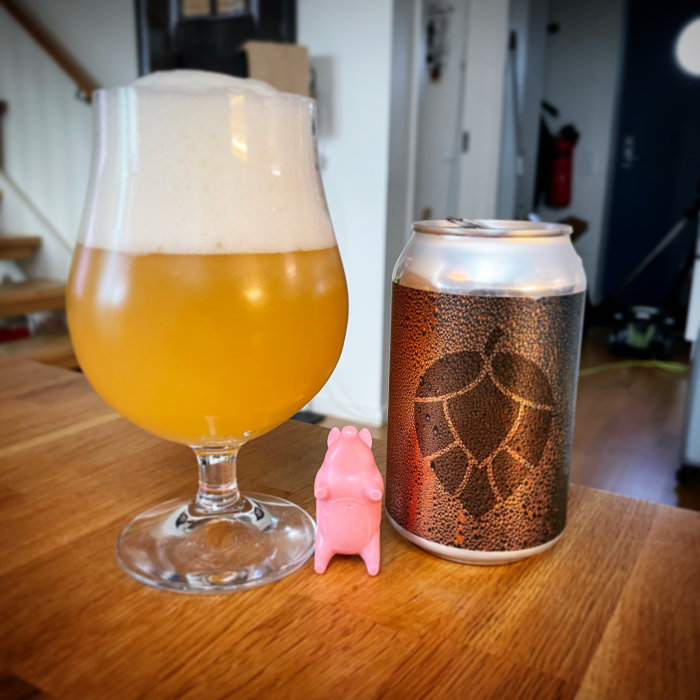 Ölglas fyllt med skummande öl bredvid en burk med mönster och en liten rosa figur på ett träbord.
