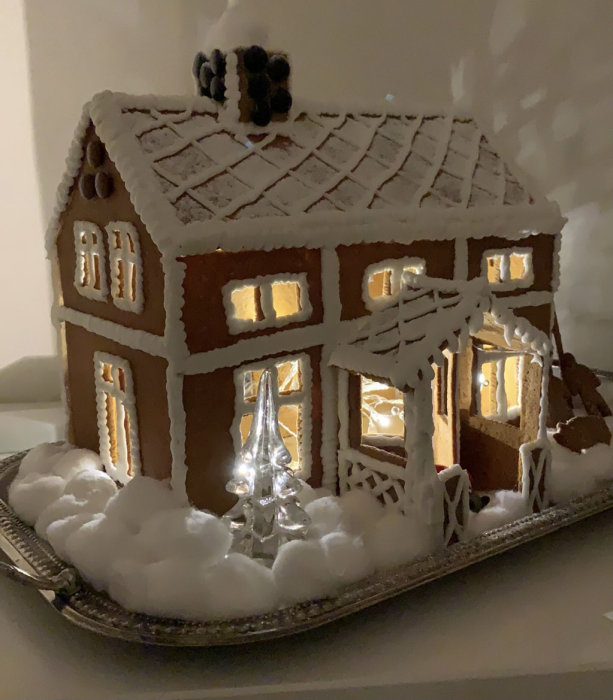 Ett detaljerat pepparkakshus på ett fat, dekorerat med kristyr och fejkad snö, belyst inifrån och en glasgran framför.