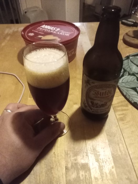 Hand håller ett glas med mörk öl bredvid en ölflaska på ett köksbord.