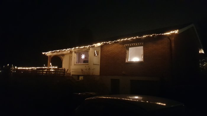 Ett hus i mörkret med tänd julbelysning längs takkanten och en lysande stjärna i fönstret.