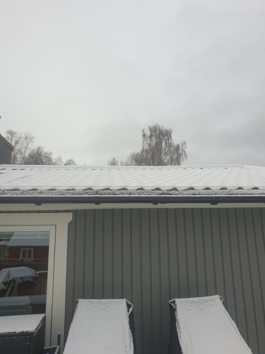 Snötäckt tegelprofil på plåttak ovanför en terrass med snöklädda utemöbler.