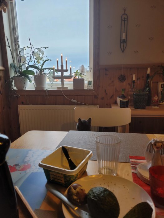 Kök med frukostbord, en katt som tittar in genom fönstret och tända ljus på fönsterbrädan.