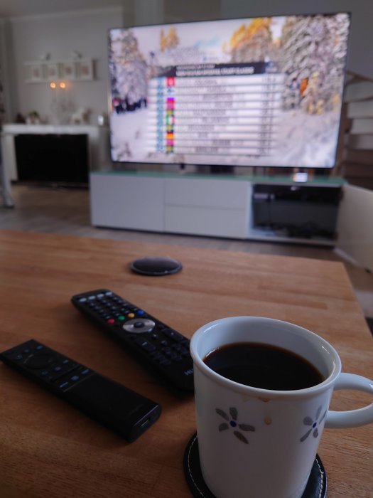 Vardagsrum med en kopp kaffe på bord framför suddig TV med text och grafik på skärmen, fjärrkontroller syns också.