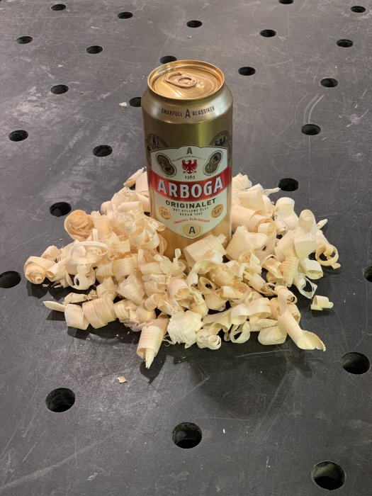 En burk Arboga Julöl omgiven av träspån på ett svart verktygsbord med hål.