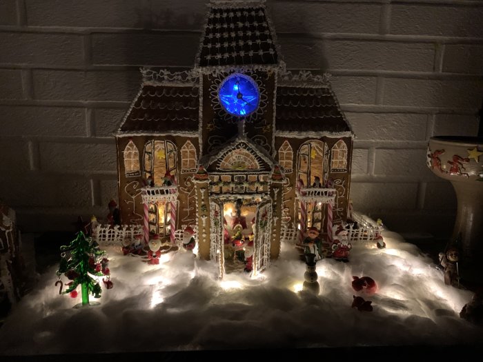 Intrikat pepparkakshus med belysning omgiven av juldekorationer och konstgjord snö.