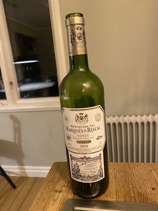 Flaska med Marques de Riscal Rioja Reserva 2016 på ett träbord mot bakgrund av ett rum.