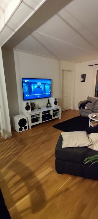 Nyrenoverat vardagsrum med trägolv och en vit TV-möbel med högtalare, inrett med grå soffa och svart fotpall.