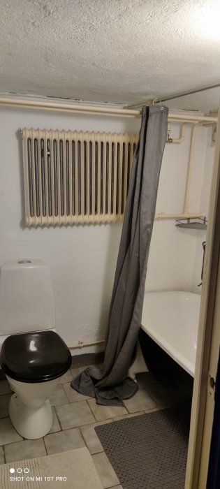 Gammalt badrum med svart toalettstol, vit badkar och duschdraperi på golvet, innan renovering.