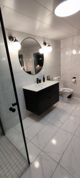 Nyligen färdigställt badrum med vit marmormönstrad kakel, svart handfatsskåp, rund spegel och duschhörna.
