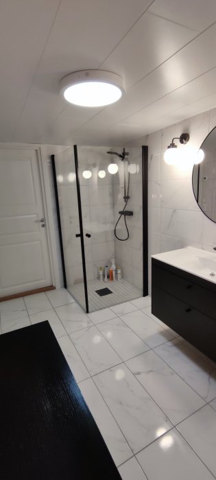 Renoverat badrum med marmormönstrade kakelplattor, duschhörna med svart inramning och vit badrumsmöbel.