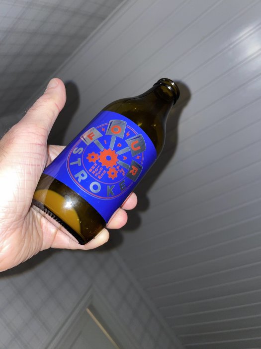 Hand håller en ölflaska med etikett "Flavour Stroke" mot en grå vägg.