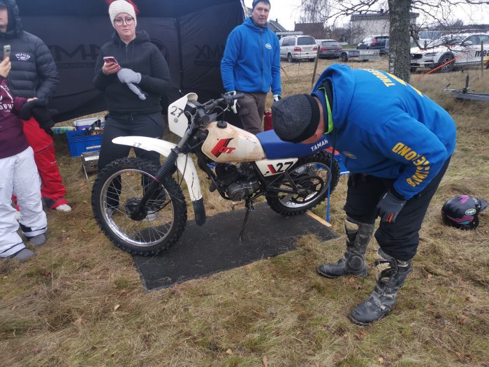 Deltagare i blå jacka justerar vintage motorcykel L27 vid stubbrace, betraktare i bakgrunden.