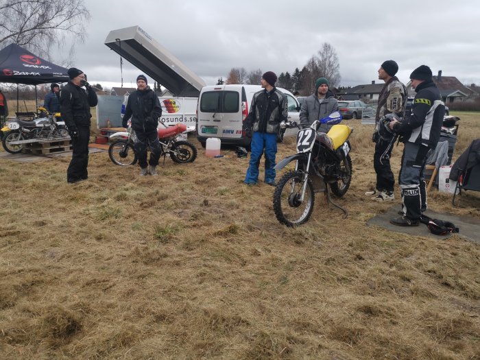 Deltagare samlade vid klassisk motorcykel med nummer 22 vid ett stubbrace, med utrustning och andra motorcyklar i bakgrunden.