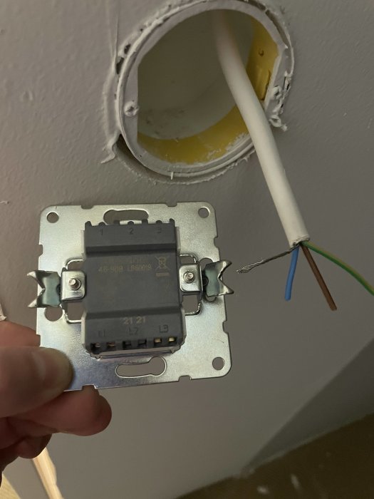 Gammal strömbrytare på vägg med ut stickande elektriska kablar i brunt, blått och grönt vid en öppen väggdosering.