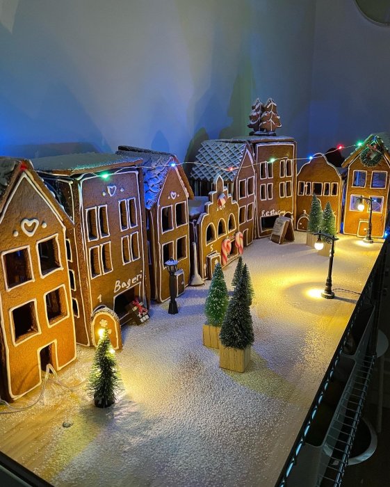 Pepparkakshus som utgör en julig by med julbelysning och små granar på ett snötäckt underlag.