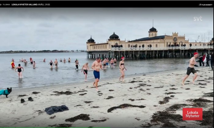 Människor badar i havet vid Varbergs kallbadhus en kylig dag, grå himmel, med klädhögar på stranden.