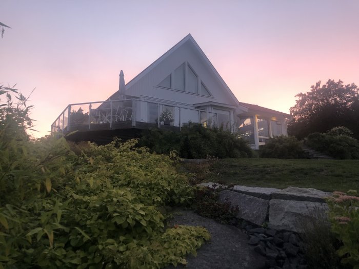 Hus med veranda under skymningshimmel, omgivet av grönska och stenläggning.