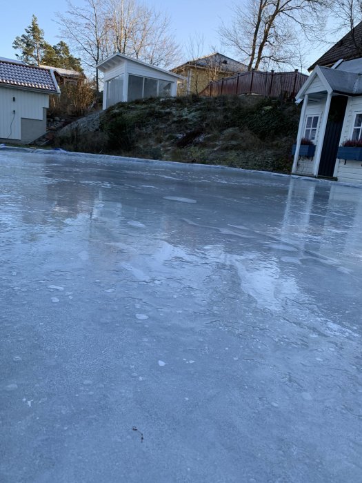 Frysen yta av vatten med omgivande hus och växtlighet i bakgrunden.