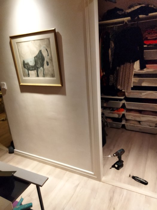 Hörn av ett rum med öppen garderobsdörr, kläder syns, en tavla på väggen och en skruvdragare på golvet.