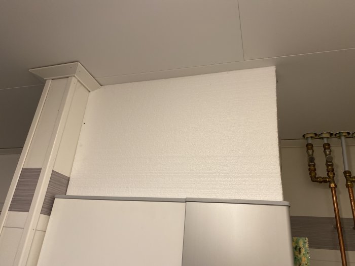 Ommålad vit vägg ovanför köksskåp med synlig maskeringstejp och rörledningar i hörnet.