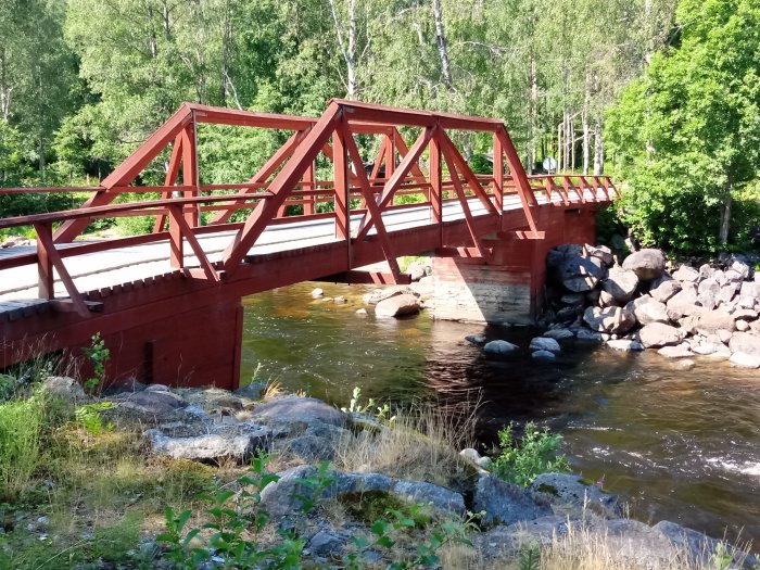 Röd träbro med fackverkskonstruktion och triangelformade räcken i en grönskande miljö i Ångermanland.
