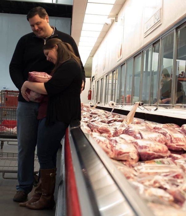 En kvinna och en man ler och kramar ett stort paket kött i en livsmedelsaffär framför en kylhylla full av köttprodukter.