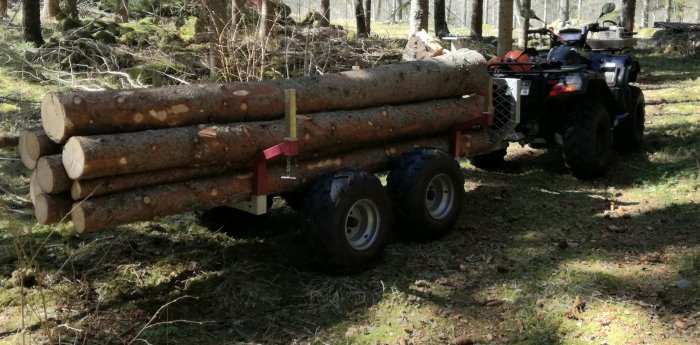 ATV med en släpvagn lastad med timmerstockar i en skogsmiljö.