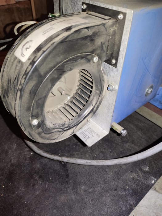 Närbild på en dammig centrifugalfläkt kopplad till en blå behållare, med en synlig etikett och elsladd.