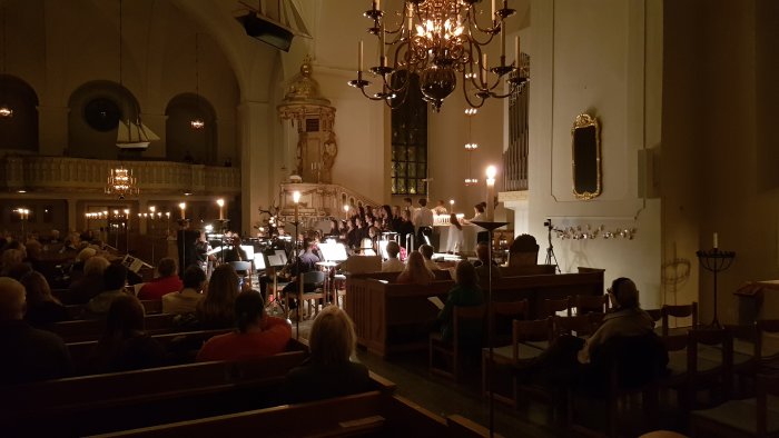 Orkester och kör framför en publik i en kyrka med tända ljuskronor och stämningsfull belysning.