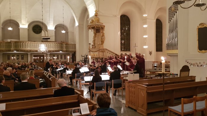 En kör och orkester framför musik framför en församling i en kyrka med vackra valv och ljuskronor.